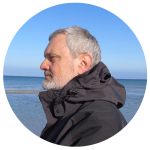 A man with a gray beard stands in profile on a beach, wearing a dark jacket. The ocean and a clear blue sky set the serene backdrop, reminiscent of Movers Brisbane guiding you effortlessly to new horizons.