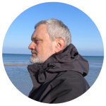 A man with a gray beard stands in profile on a beach, wearing a dark jacket. The ocean and a clear blue sky set the serene backdrop, reminiscent of Movers Brisbane guiding you effortlessly to new horizons.