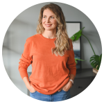 Woman with long hair, wearing an orange sweater, stands with hands in pockets, looking to the side. A plant and framed picture are in the background. Her contemplative gaze suggests thoughts of a fresh start, perhaps spurred by recent experiences with movers in Brisbane.