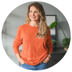 Woman with long hair, wearing an orange sweater, stands with hands in pockets, looking to the side. A plant and framed picture are in the background. Her contemplative gaze suggests thoughts of a fresh start, perhaps spurred by recent experiences with movers in Brisbane.